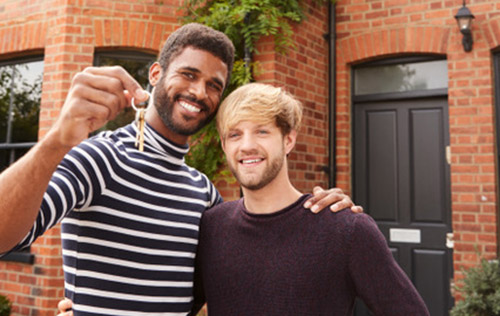 Portrait Of Excited Gay Male Couple Standing Outside New Home Holding Keys On Moving Day Together